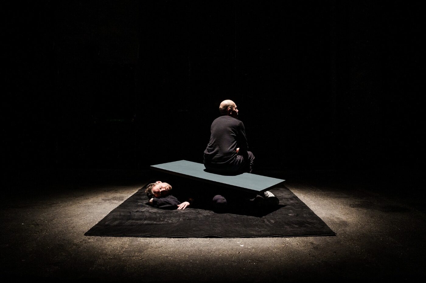 Kenneth Kvarnström sitter på en bräda som ligger ovanpå lutisten Jonas Nordberg i spotlight nedslag i svart black box