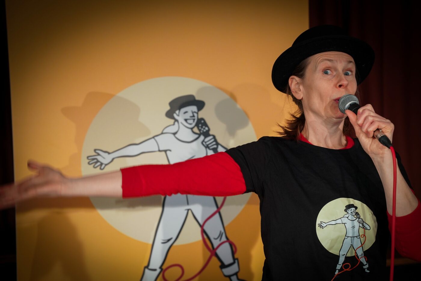 Michaela står framför en ritad bild av henne själv med höger arm rakt ut och håller i en mikrofon. Hon har en svart hatt på huvudet samt en röd tröja med en svart t-shirt med tryck på sig.