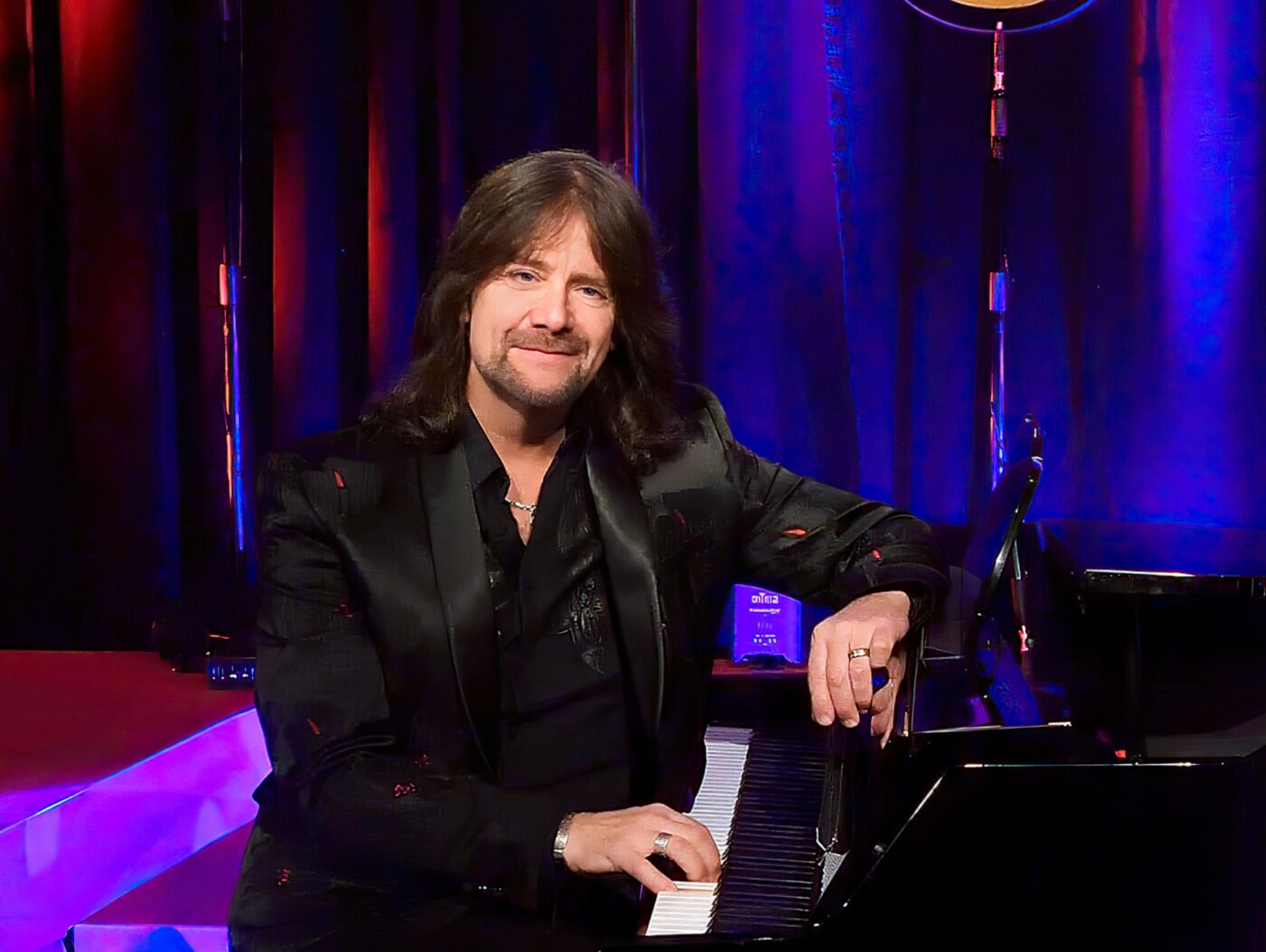 Robert Wells sitter vid flugeln med ena handen på pianotangenterna och ler mot oss. Han har långt hår och svart kostym.