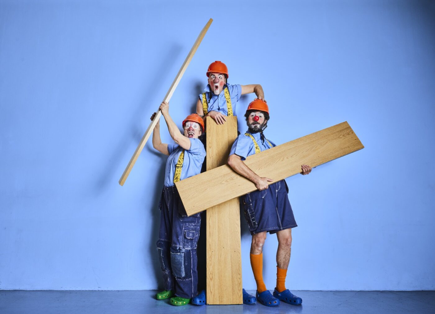 Tre clowner och skådespelare står med tre stora träplankor. De har orangea hjälmar, röd lösnäsa och scenkostym i blå toner