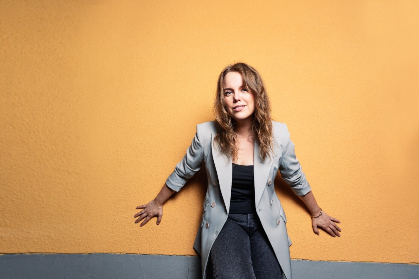 Kvinna i mellanlångt år och grå lång kavaj står lutad mot en gul vägg