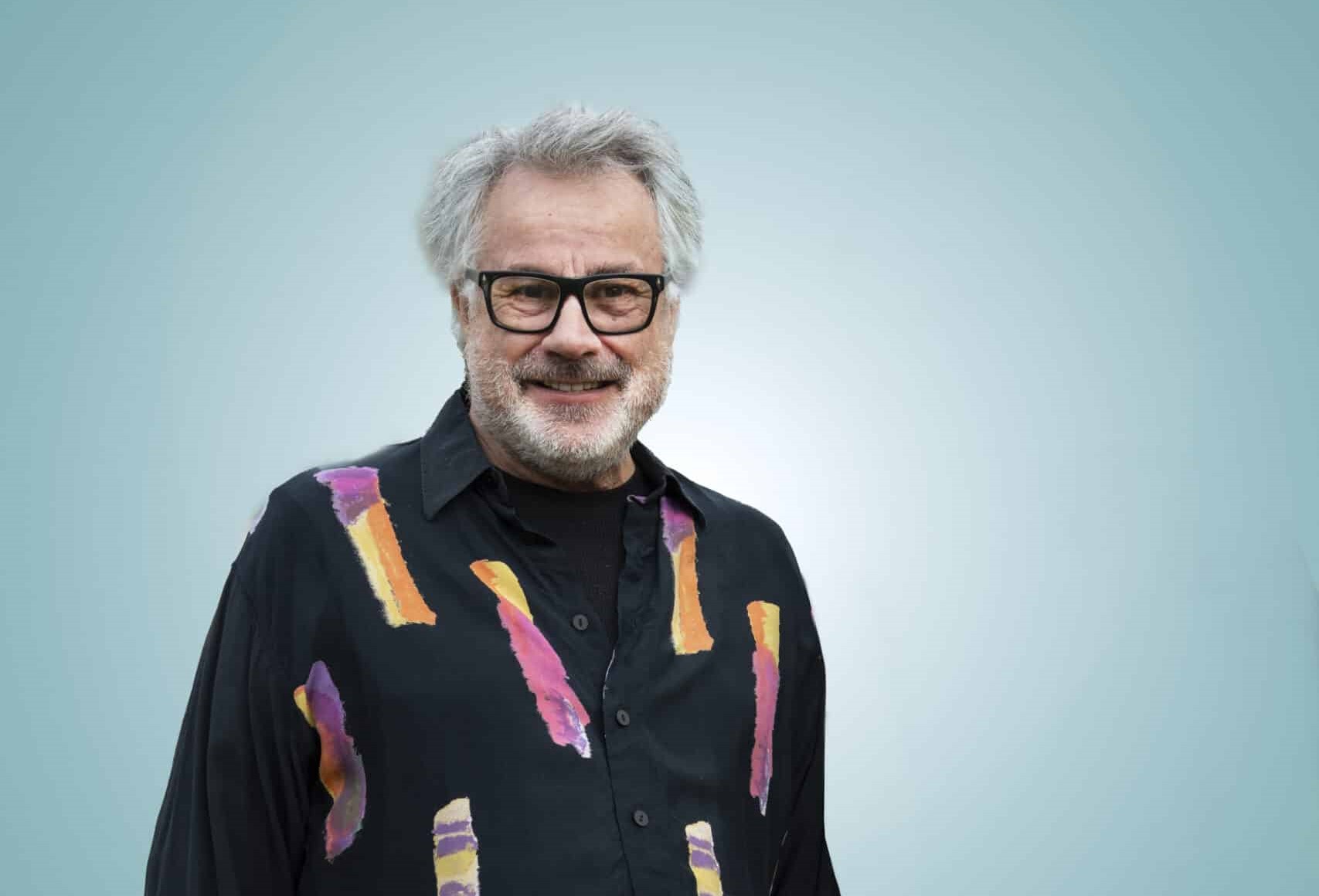 Leende man i gråvitt hår och skägg med svartbågade glasögon iklädd en svart skjorta med färgglatt mönster.