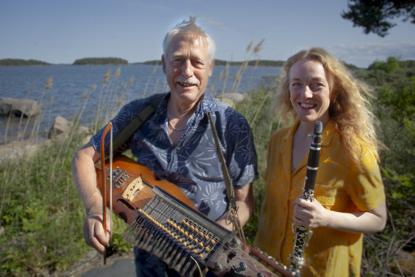 Man i blå skjorta med nyckelharpa och kvinna i gul klänning med klarinett står i solen framför landskap med sjö och gräs