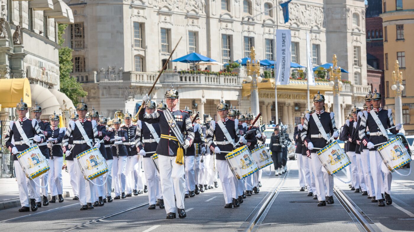 En militärorkester i uniform marscherar längs en stadsgata, ledda av en tamburmajor. Musikerna har mörka jackor, vita byxor, höga stövlar och guldfärgade hjälmar. Flera av dem spelar på stora trummor. I bakgrunden syns byggnaden till Kungliga Dramatiska Teatern i Stockholm, med dess utsmyckade fasad och blå parasoller på balkongen. Andra pampiga byggnader och människor syns längs gatan.