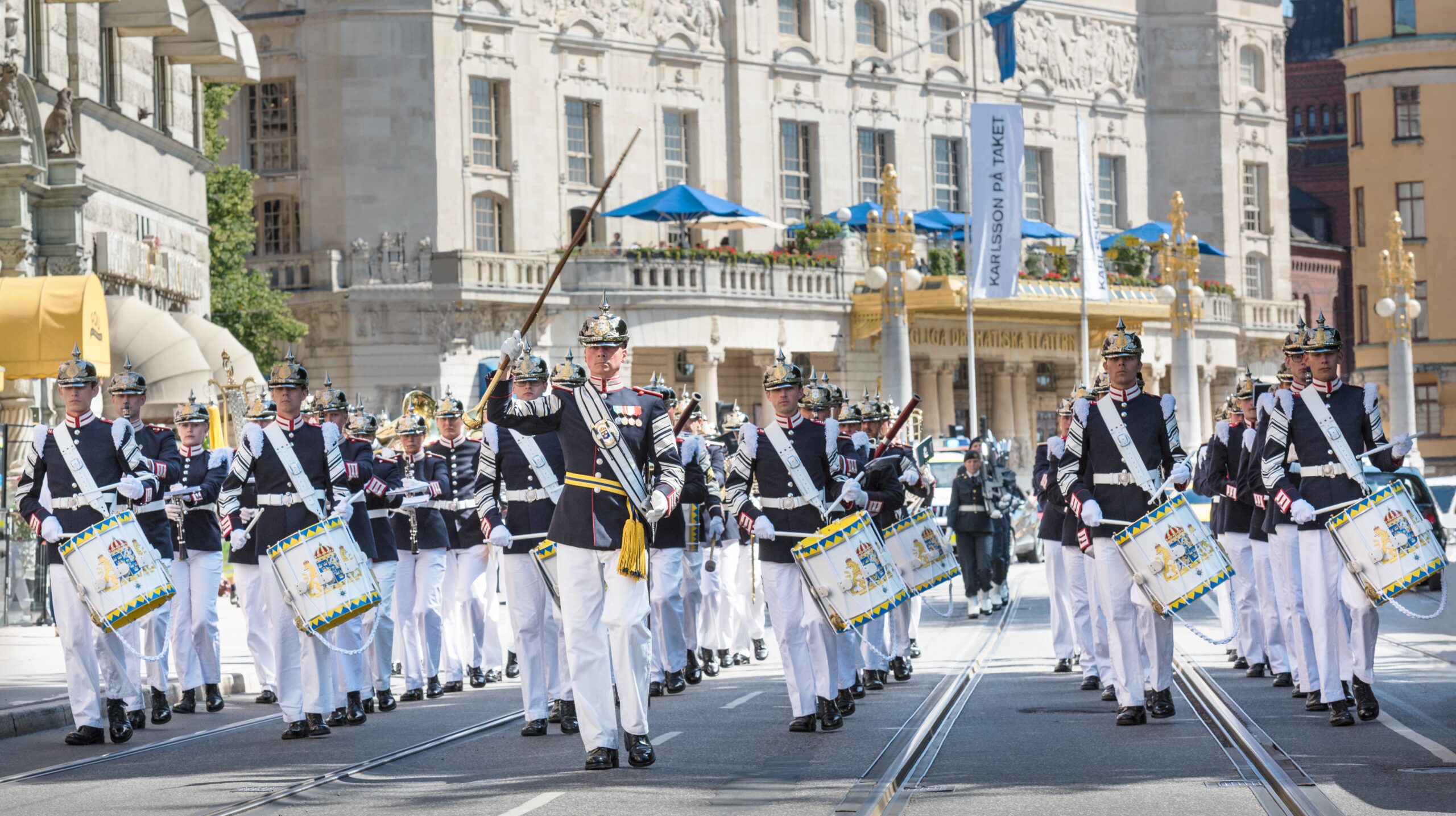 En militärorkester i uniform marscherar längs en stadsgata, ledda av en tamburmajor. Musikerna har mörka jackor, vita byxor, höga stövlar och guldfärgade hjälmar. Flera av dem spelar på stora trummor. I bakgrunden syns byggnaden till Kungliga Dramatiska Teatern i Stockholm, med dess utsmyckade fasad och blå parasoller på balkongen. Andra pampiga byggnader och människor syns längs gatan.