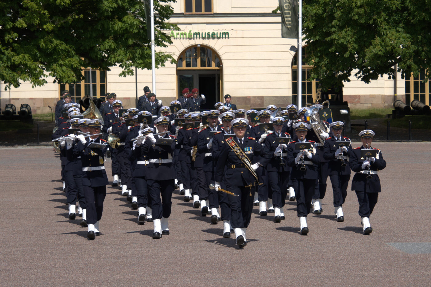 En uniformerad militärorkester marscherar och spelar instrument utanför Armémuseum. Orkestern leds av en tamburmajor och består av musiker i mörka uniformer, vita handskar och vita mössor. I bakgrunden syns museets byggnad och några träd.