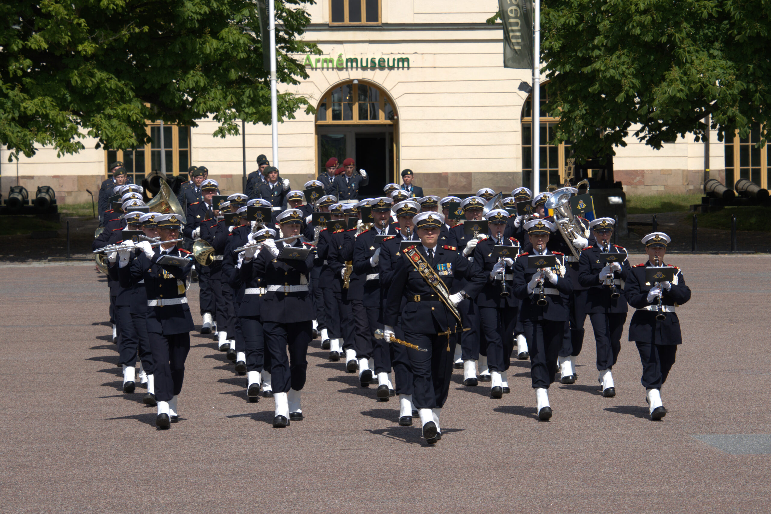 En uniformerad militärorkester marscherar och spelar instrument utanför Armémuseum. Orkestern leds av en tamburmajor och består av musiker i mörka uniformer, vita handskar och vita mössor. I bakgrunden syns museets byggnad och några träd.