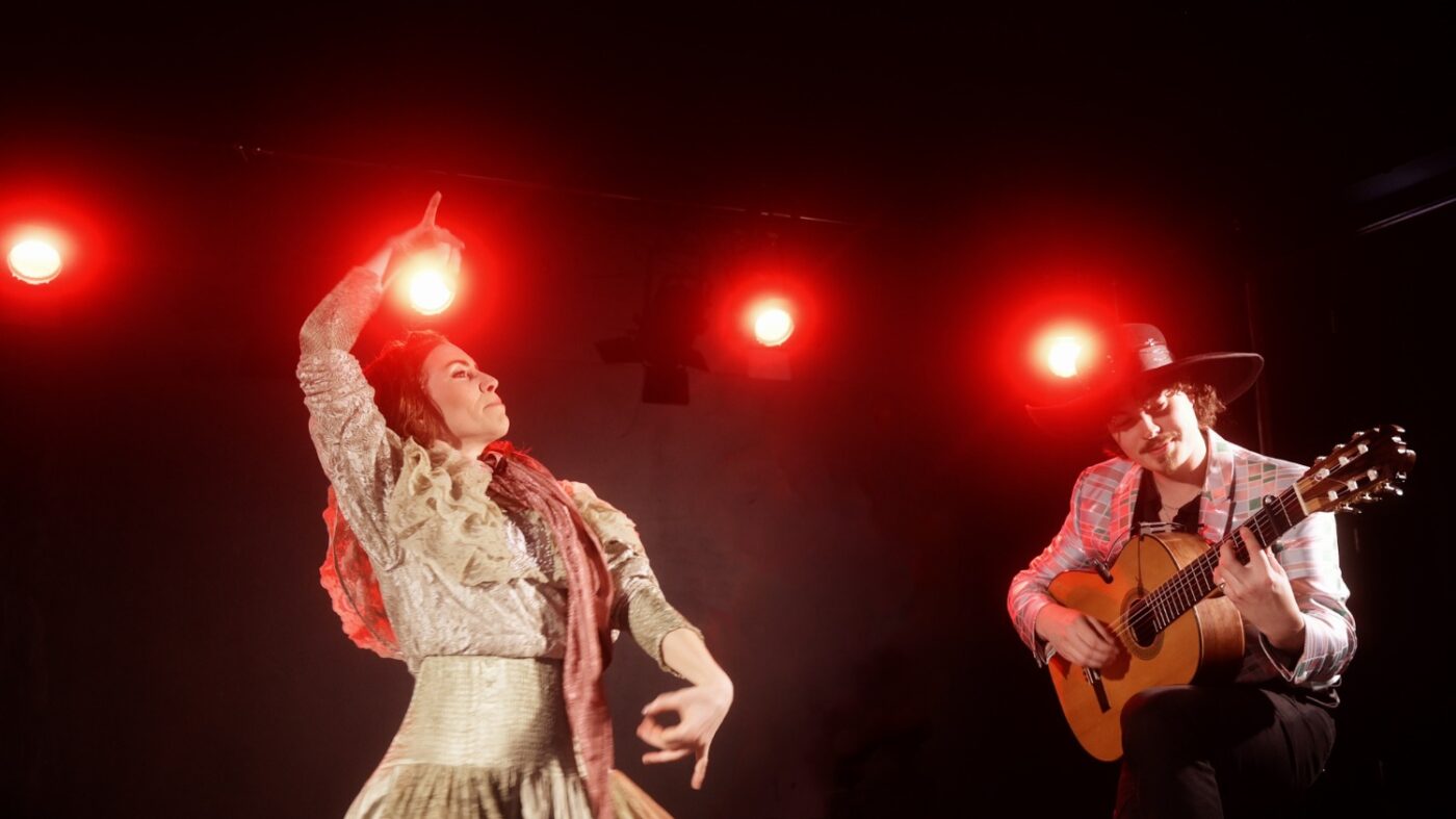 flamencodansare i vit klänning och en gitarrist i hatt på en scen med röda spotlights i bakgrunden