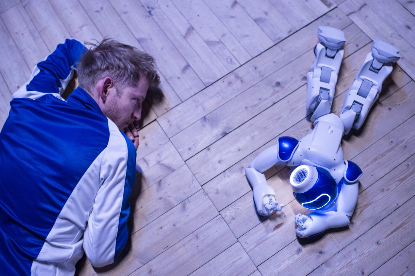 En man i blåvit tröja ligger på mage på ett trägolv och tittar på en liten robot som också ligger ned på mage.